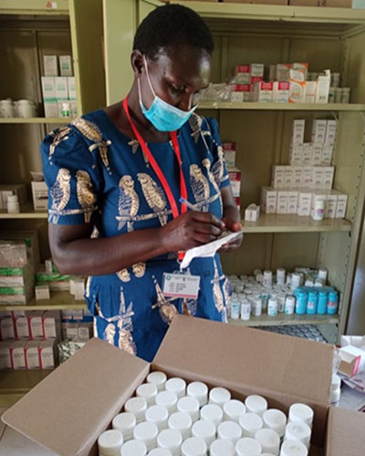 A person wearing a blue mask inspects a box of medication and writes on a piece of paper.
