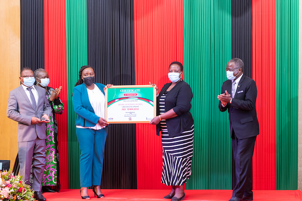Two people pose for a photo with a certificate while surrounded by others clapping.