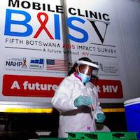 A person wearing protective equipment stands in front of a mobile clinic BAIS V sign