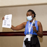 A person wearing a face mask holds a microphone and a piece of paper.