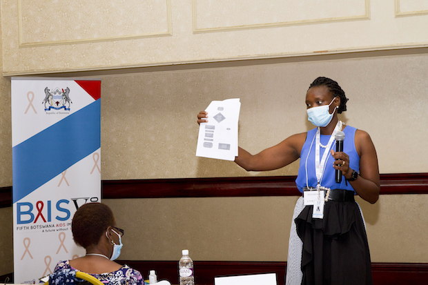 A woman holds a packet of papers and microphone while standing and speaking in front of a person.