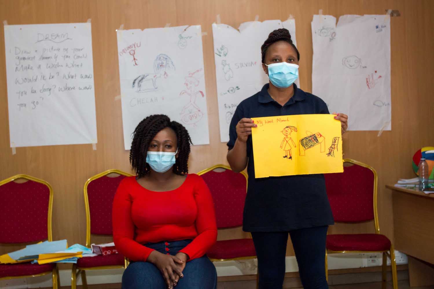 One woman wearing a red shirt and blue face mask is sitting, while another woman wearing a black shirt and blue face mask stands holding a yellow paper with drawings on it.