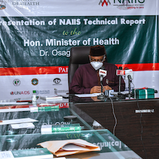 A person wearing a face mask sits at a conference table with microphones in front of him.