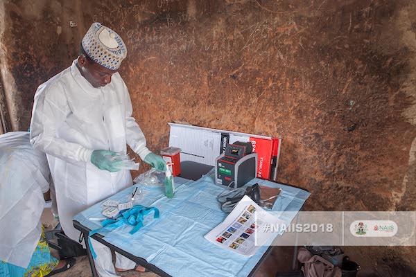 A person wearing a white gown and green gloves looks at medical supplies.