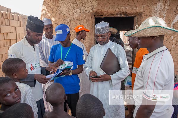 A group of field workers look at their phones and papers while a group of children stand in front of them.