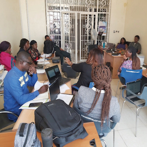 A group of people sit around computers and desks in a discussion.