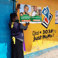 A person wearing a face mask holds up a poster on a colorful yellow wall with art painted on it.